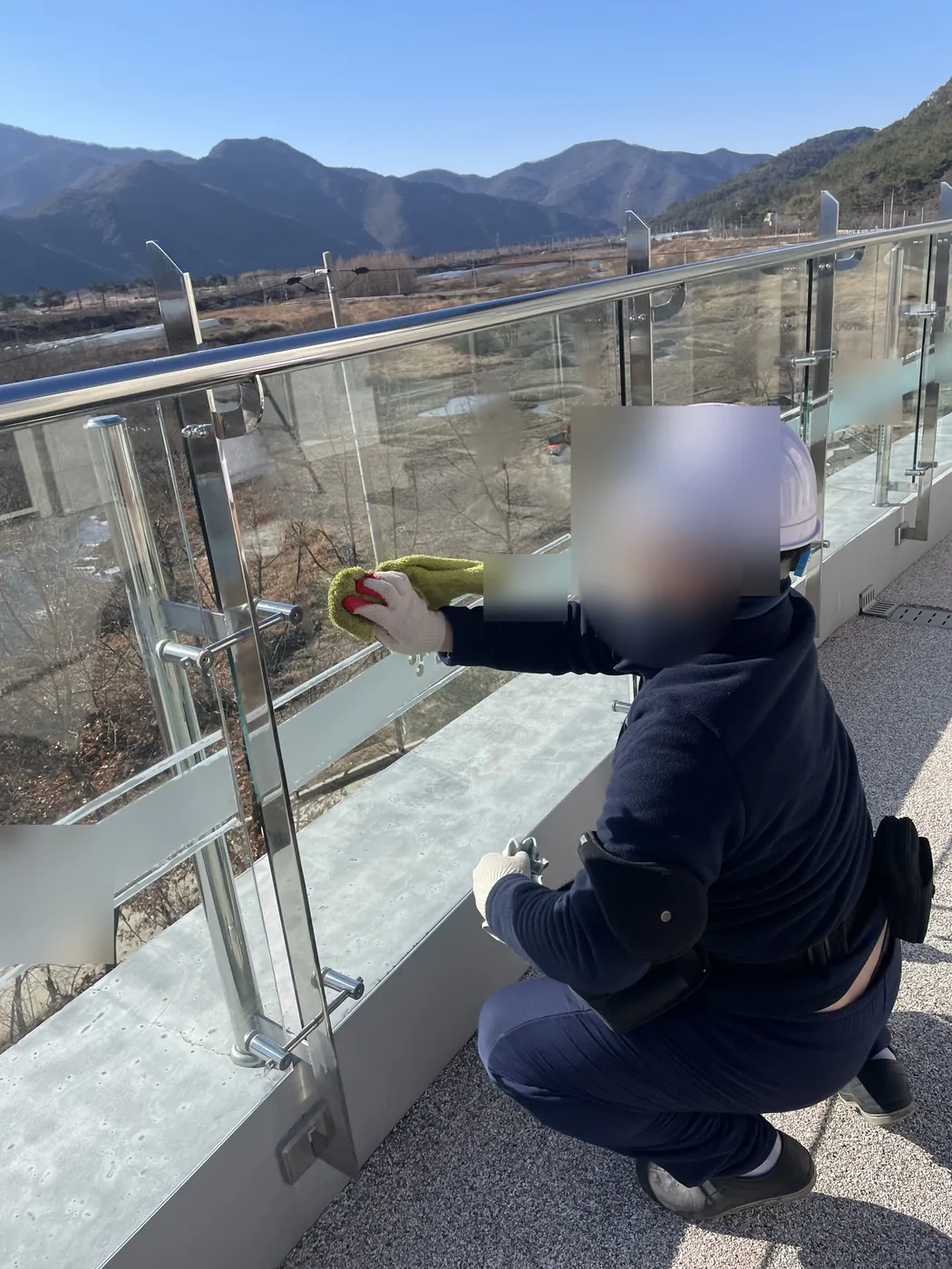 옥상 데크에서 안전모를 쓴 작업자가 쪼그려 앉아 유리 난간을 천으로 닦는 모습