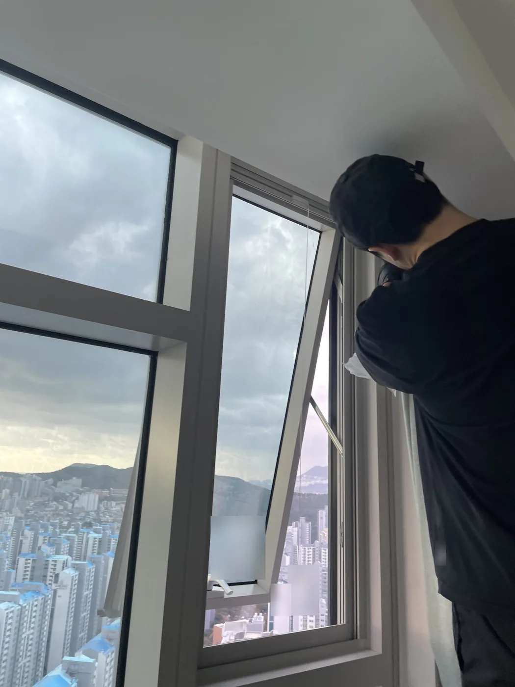 부산 남구 고층 아파트에서 작업자가 안쪽으로 젖혀 연 창 옆에 서서 창틀 가장자리를 닦는 뒷모습