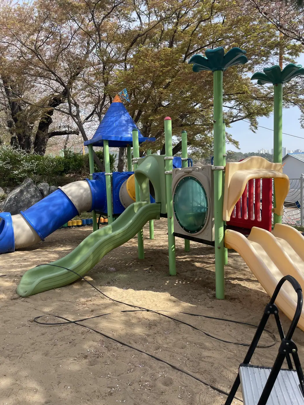 나무 아래 모래밭 위에 튜브 미끄럼틀과 원뿔 지붕, 여러 갈래 미끄럼틀을 갖춘 복합 놀이대 전경