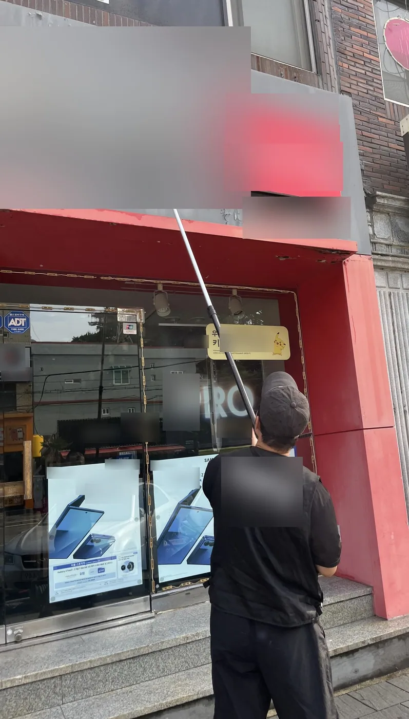 울산 동구 휴대폰 매장 입구 위 회색 간판 판넬을 작업자가 긴 폴대 끝 솔로 문질러 닦는 뒷모습