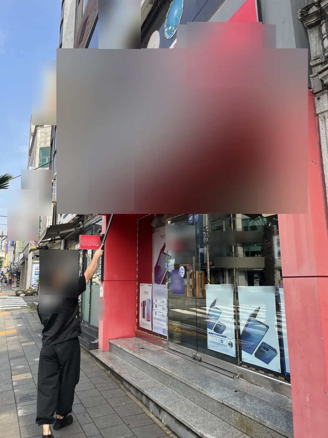 인도에 선 작업자가 폴대를 위로 뻗어 붉은 테두리 위 간판 판넬 왼쪽을 솔로 닦는 모습