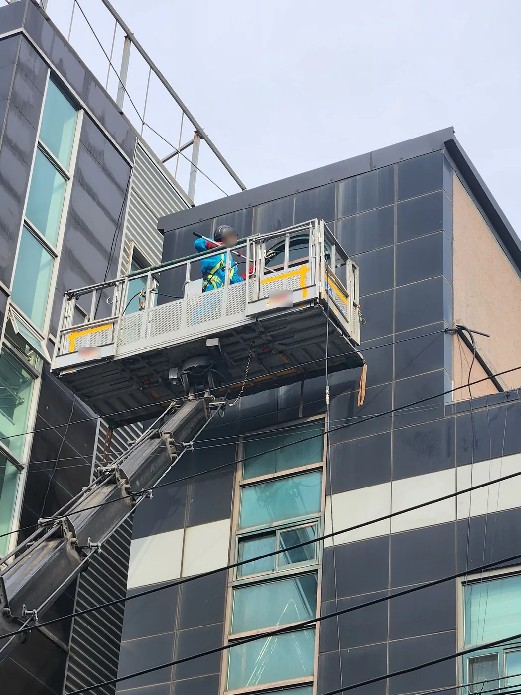 포항 상가건물 외벽 윗단에 고소작업차 바스켓을 붙이고 작업자가 긴 봉으로 세척하는 장면