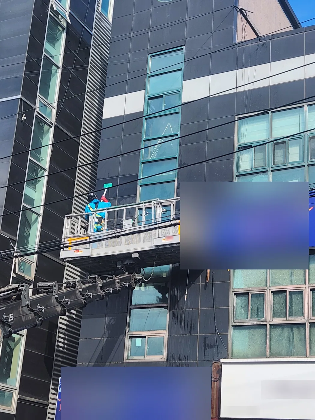 고소작업차 붐 끝 바스켓에 선 작업자가 건물 세로 창 라인을 긴 봉으로 닦는 모습