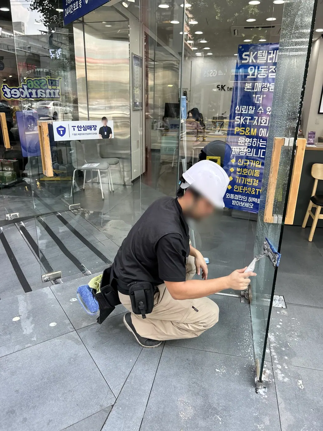 김해 통신매장 입구에서 헬멧을 쓴 작업자가 쪼그려 앉아 유리문 아랫단을 스퀴지로 걷어 내는 모습