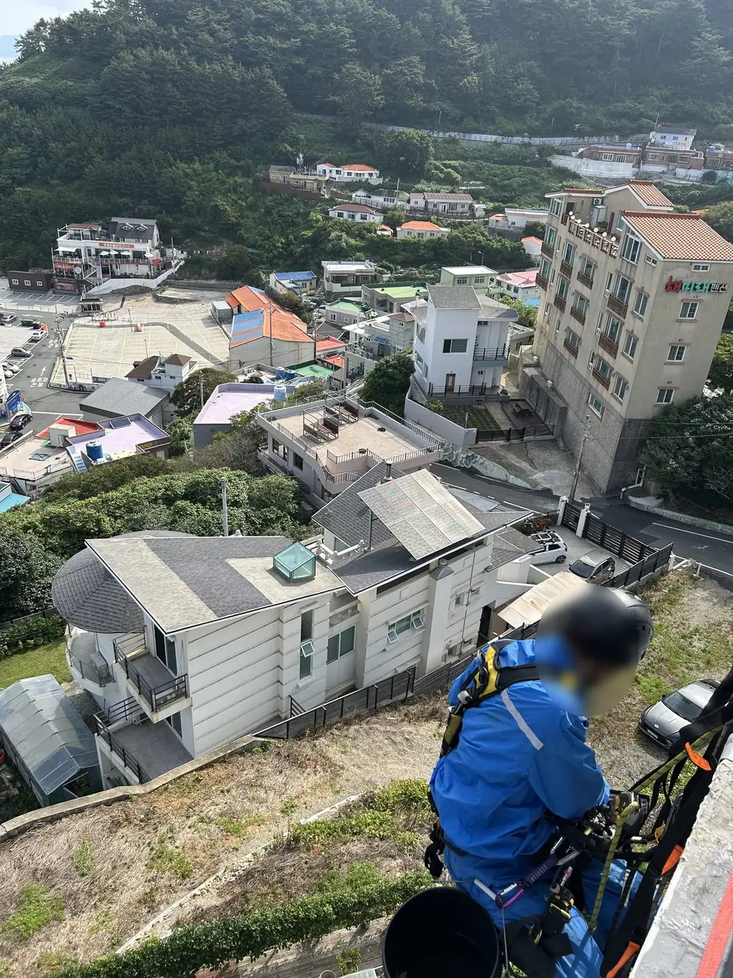거제 바닷가 카페 건물 외벽에서 헬멧과 안전대를 갖춘 작업자가 로프에 매달려 창문을 닦는 모습