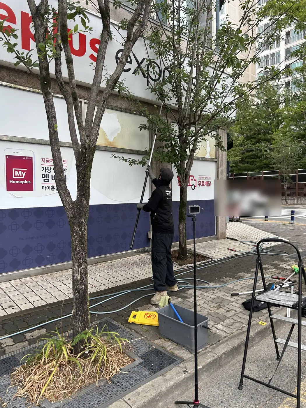 김해 대형마트 매장 앞 가로수 옆에서 작업자가 긴 연장봉 브러시로 흰 판형 간판을 닦는 모습