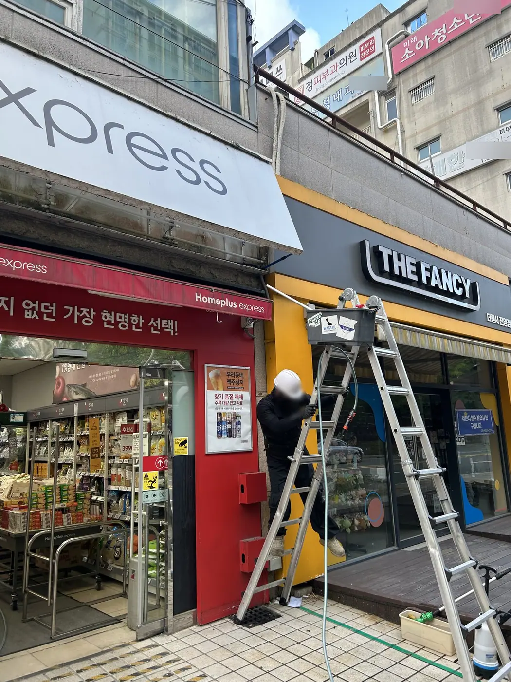 작업자가 매장 모서리 사다리 위에서 간판 아랫단과 처마를 닦는 모습