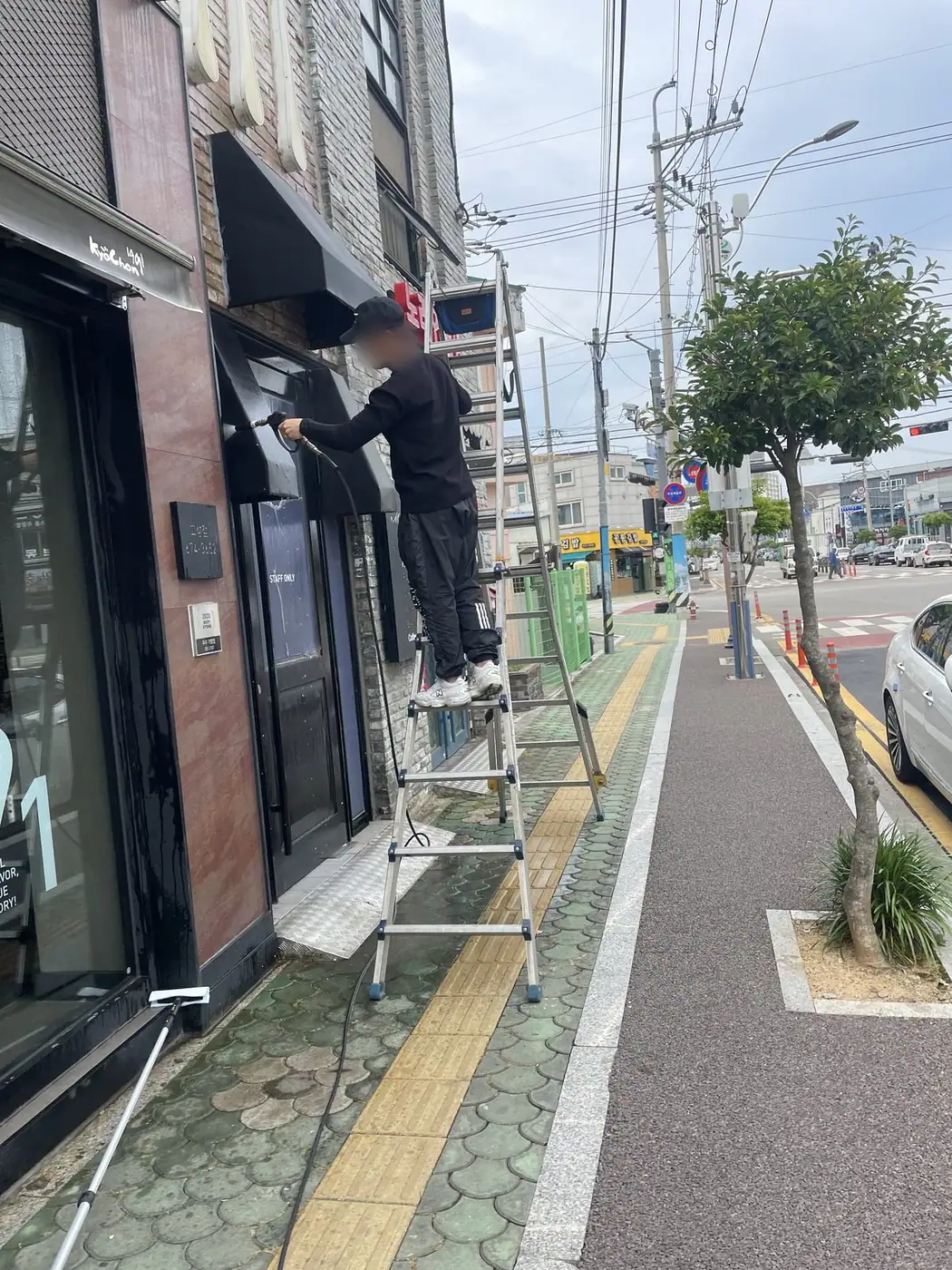 도로변 보도에 사다리를 세우고 작업자가 어닝을 세척하는 장면