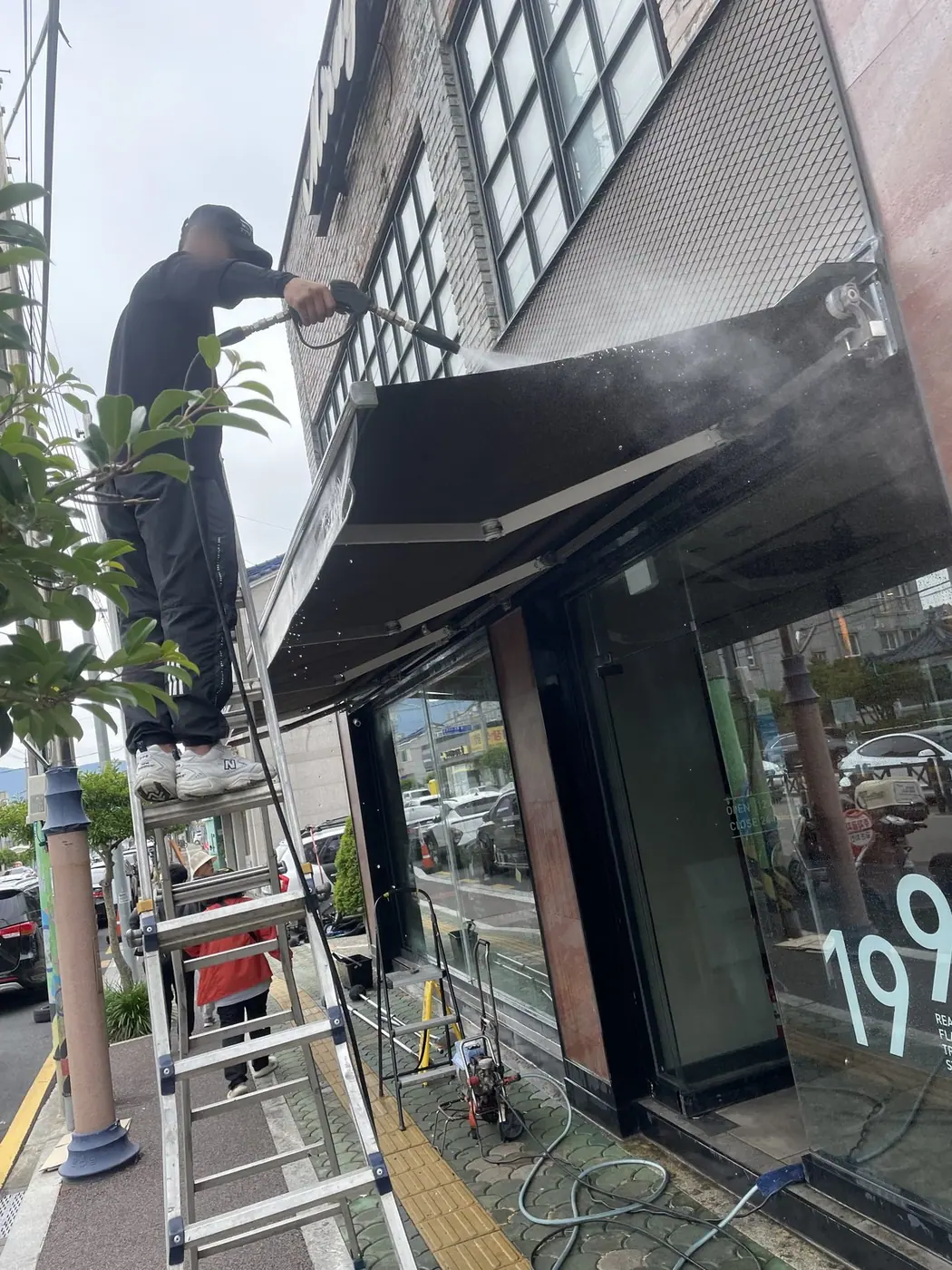 도로변 보도에 사다리를 세우고 작업자가 어닝을 세척하는 장면
