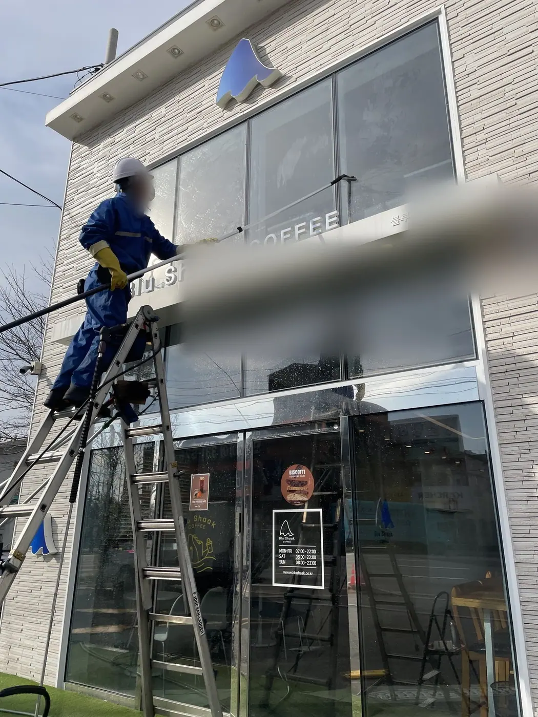 점포 전면 한쪽 끝 구간에서 작업자가 사다리에 올라 유리를 세척하는 장면