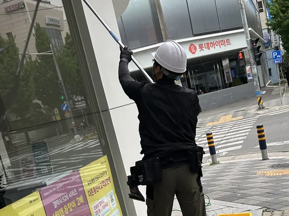 흰 안전모를 쓰고 긴 폴대로 모퉁이 매장 윗유리를 닦는 작업자